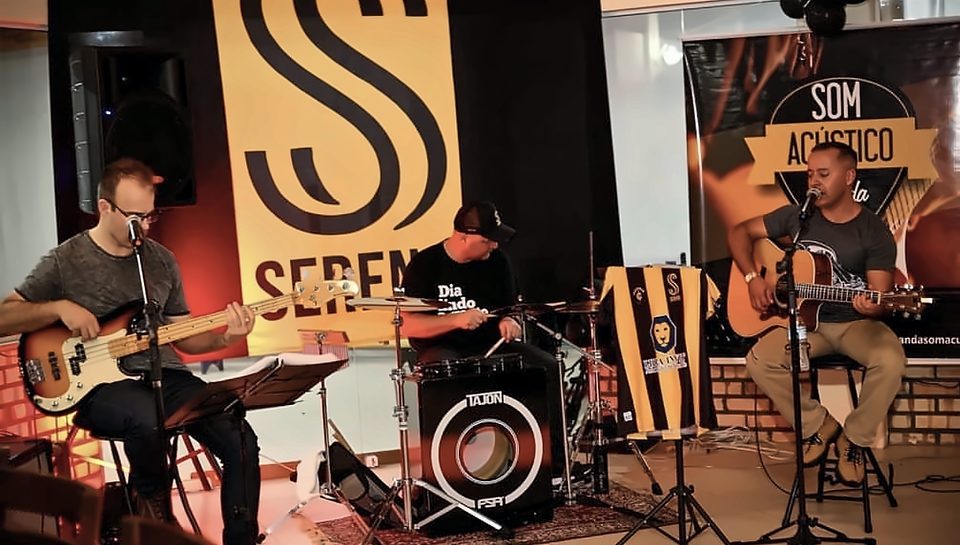 Os três integrantes se apresentando em um salão: contrabaixo à esquerda, percussão ao centro e violão à direita