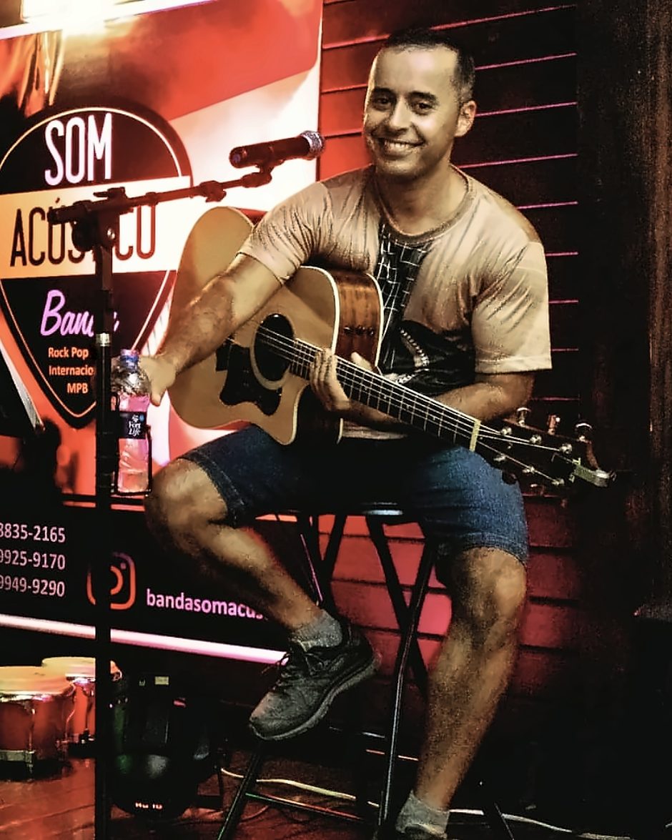 Alan Peter sorrindo, sentado com o violão em frente ao banner da banda